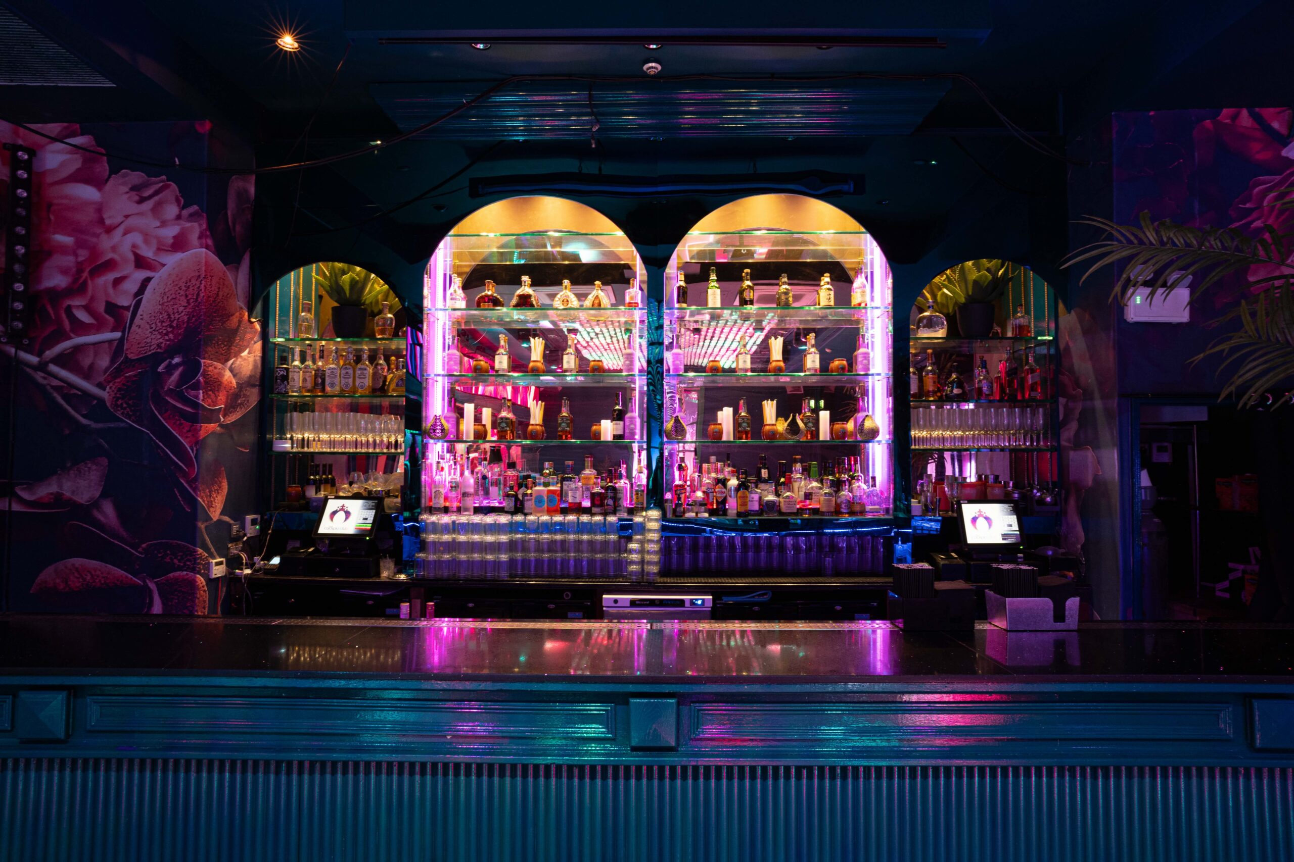 Bar with twin arched backlit shelves of bottles, neon pink lighting and turquoise counter